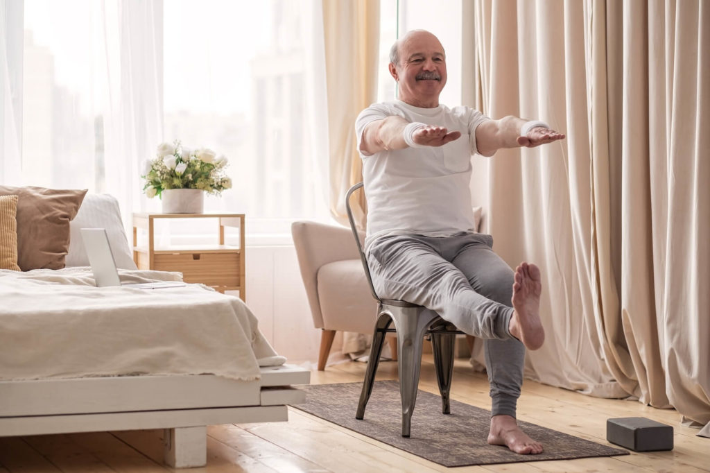 An older adult smiling during chair yoga in a sunlit bedroom to reduce their risk of falls.