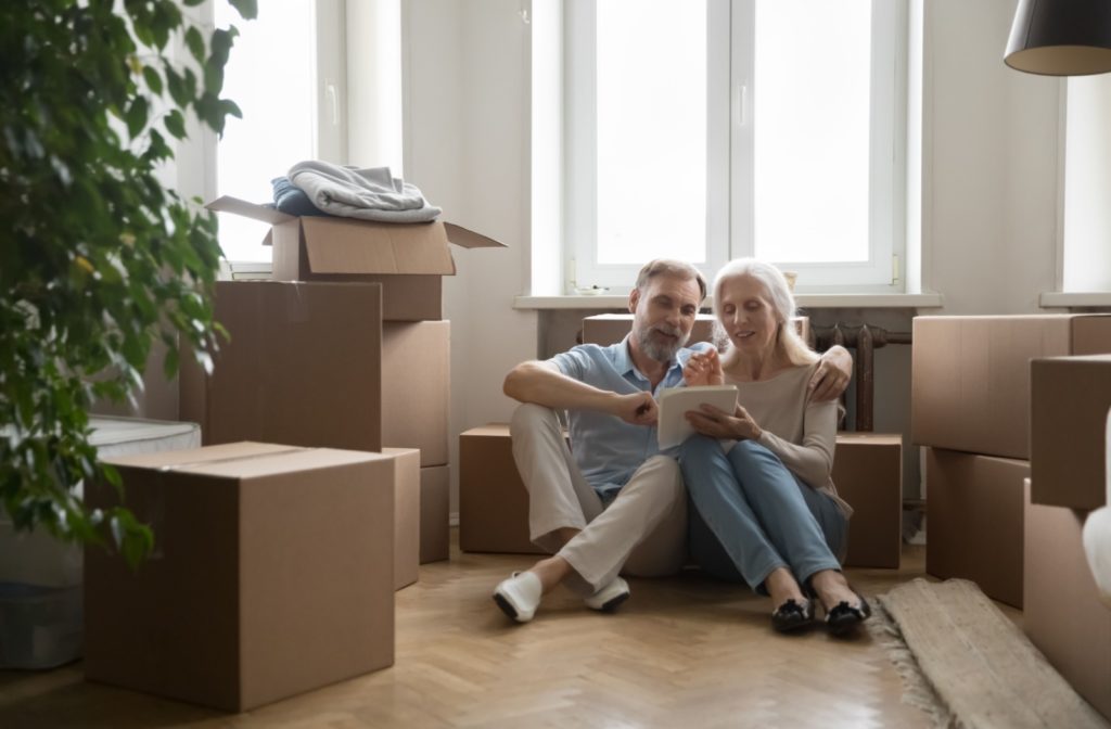 An older adult couple sitting on the floor of their home, surrounded by boxes, preparing to move to an assisted living community.