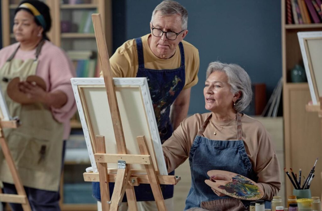 A senior sitting at an easel during a painting class provided by their senior living community gets advice from a classmate