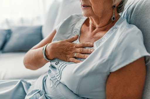 A senior woman sits on a couch with a hand on her chest.
