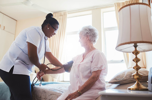 A nurse stands and takes a senior woman's blood pressure while she sits on a bed.