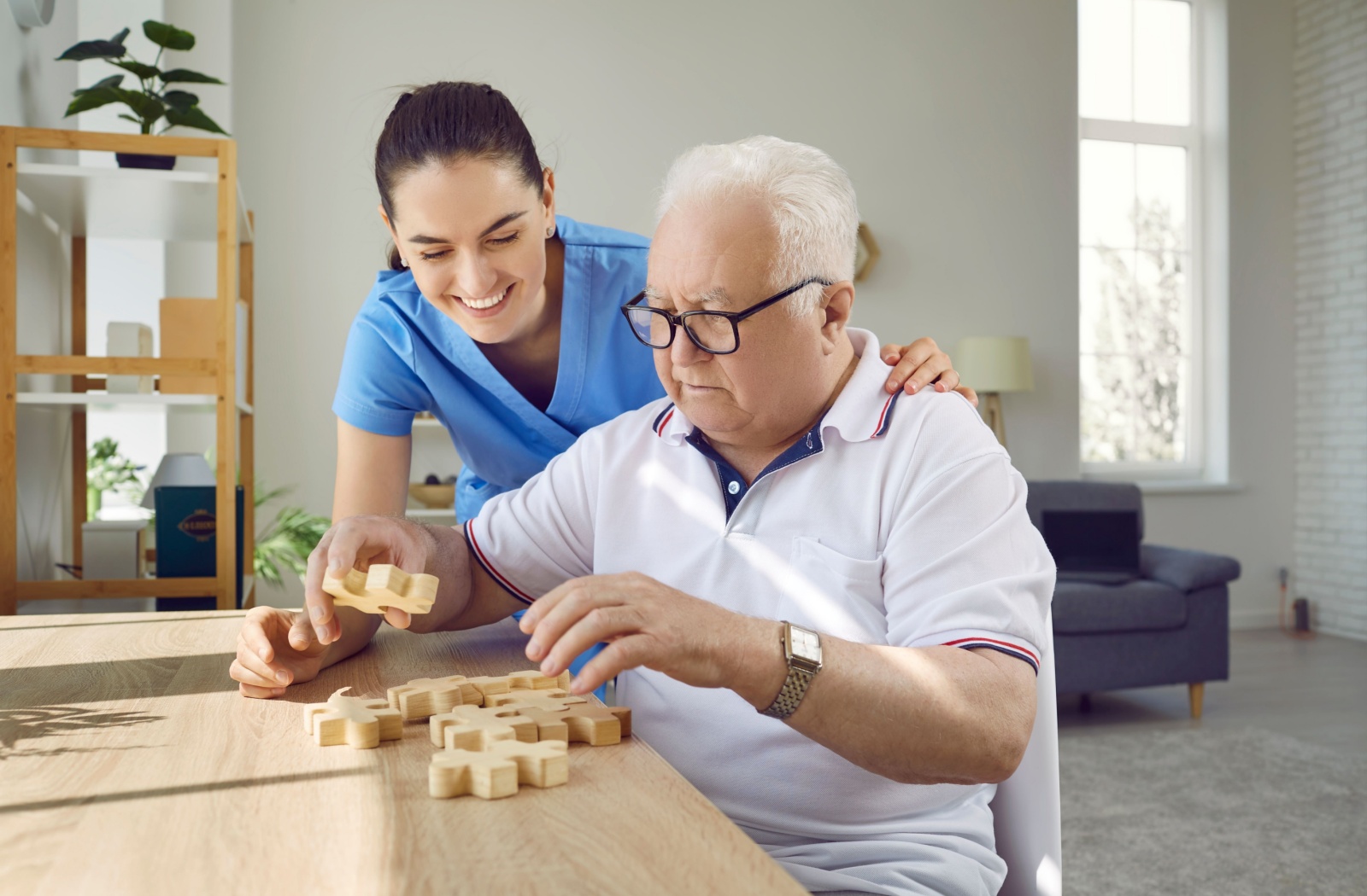 An older adult man solving a wooden jigsaw puzzle with the help of a memory care staff.