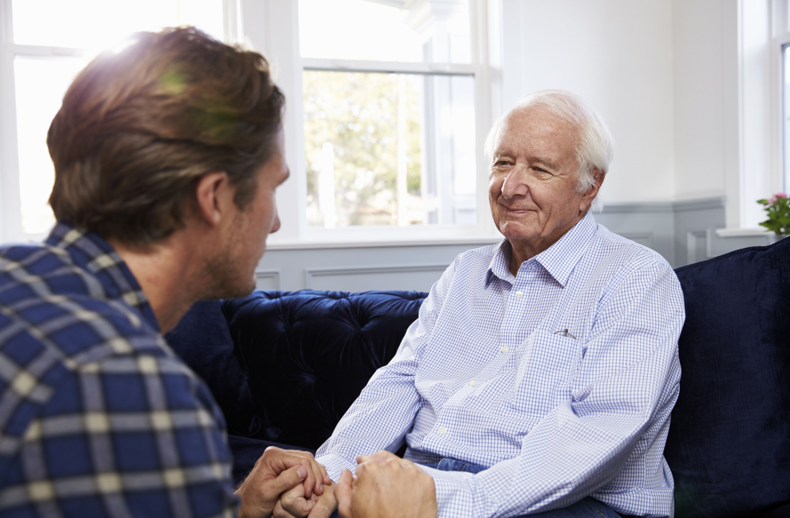 An adult child sitting in front of an older adult on a couch with dementia holding both his hands.