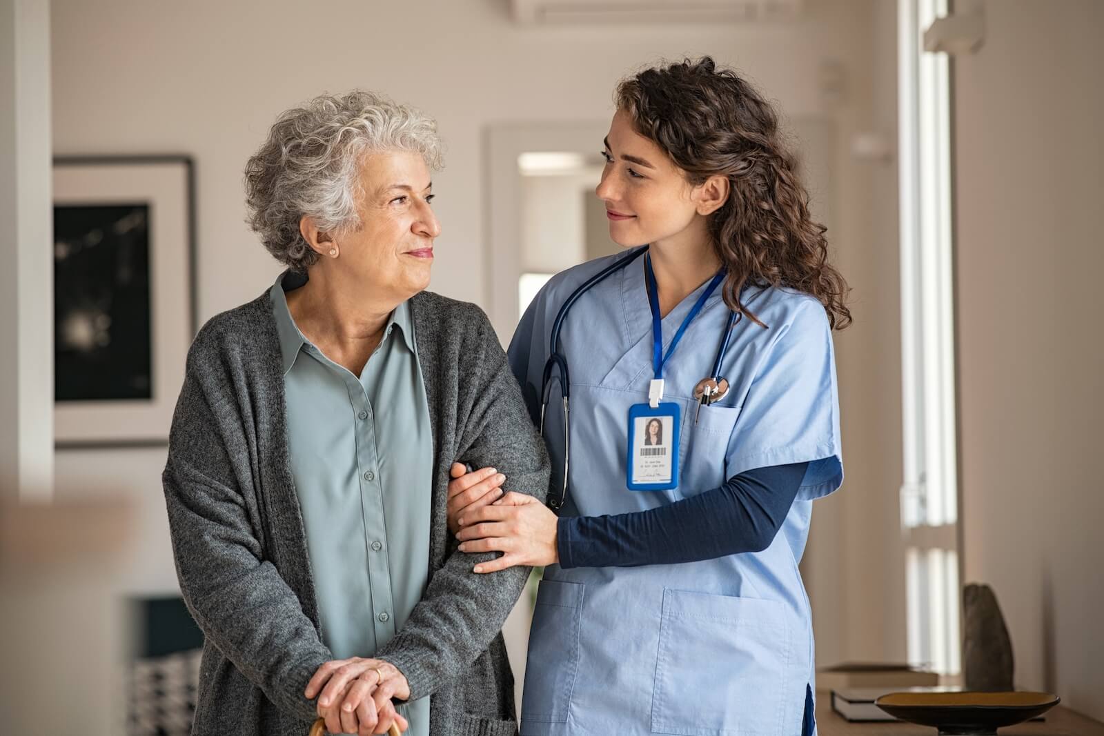 A caregiver helping an older adult walk down the hall to avoid falls while they smile at each other in appreciation