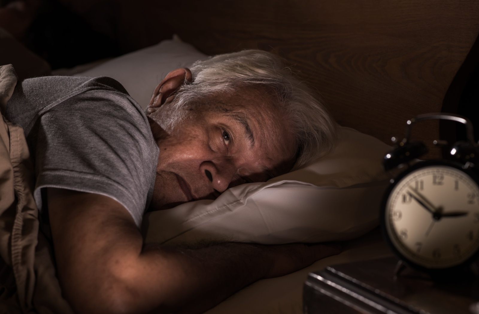 A sleeping-looking senior lying in bed next to an alarm clock.