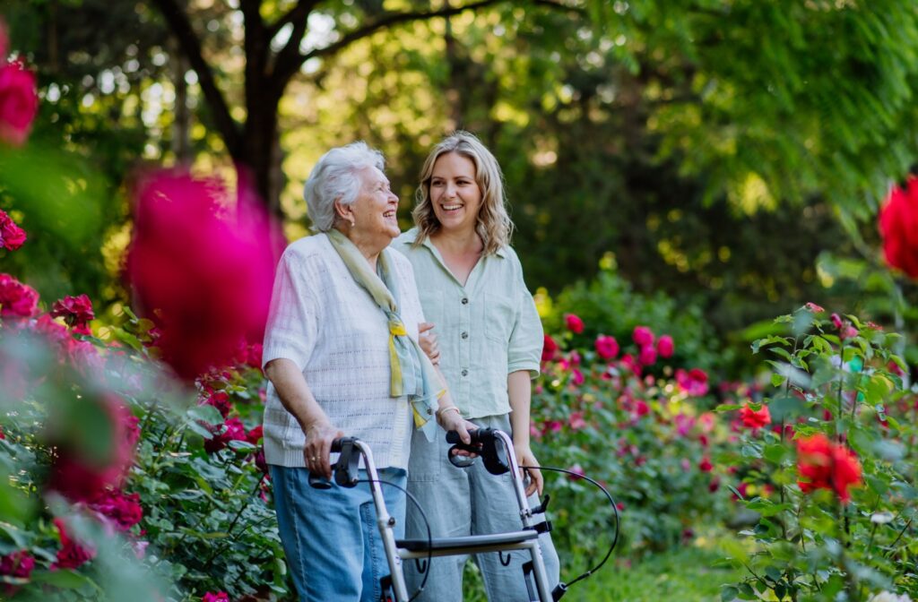 A senior woman using a walker, walking alongside her daughter outdoors.