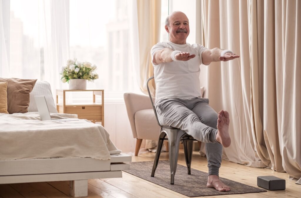 An older adult smiling while performing a seated leg raise in a sunlit room.