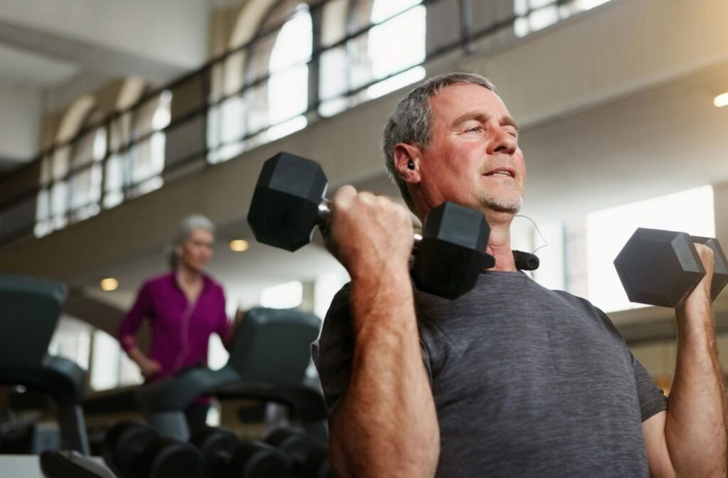 Older adult lifting dumbbells in a gym setting with focused expression and natural light coming through large windows