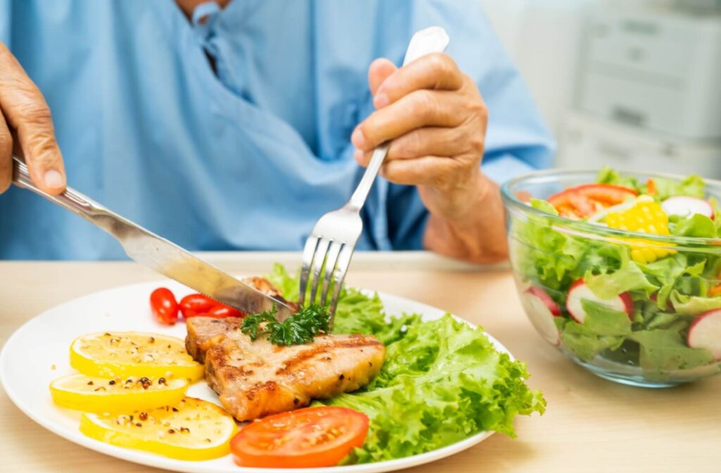 Close-up of older adult eating a protein-rich meal with grilled fish, squash, tomatoes, and a fresh green salad in a bright dining space