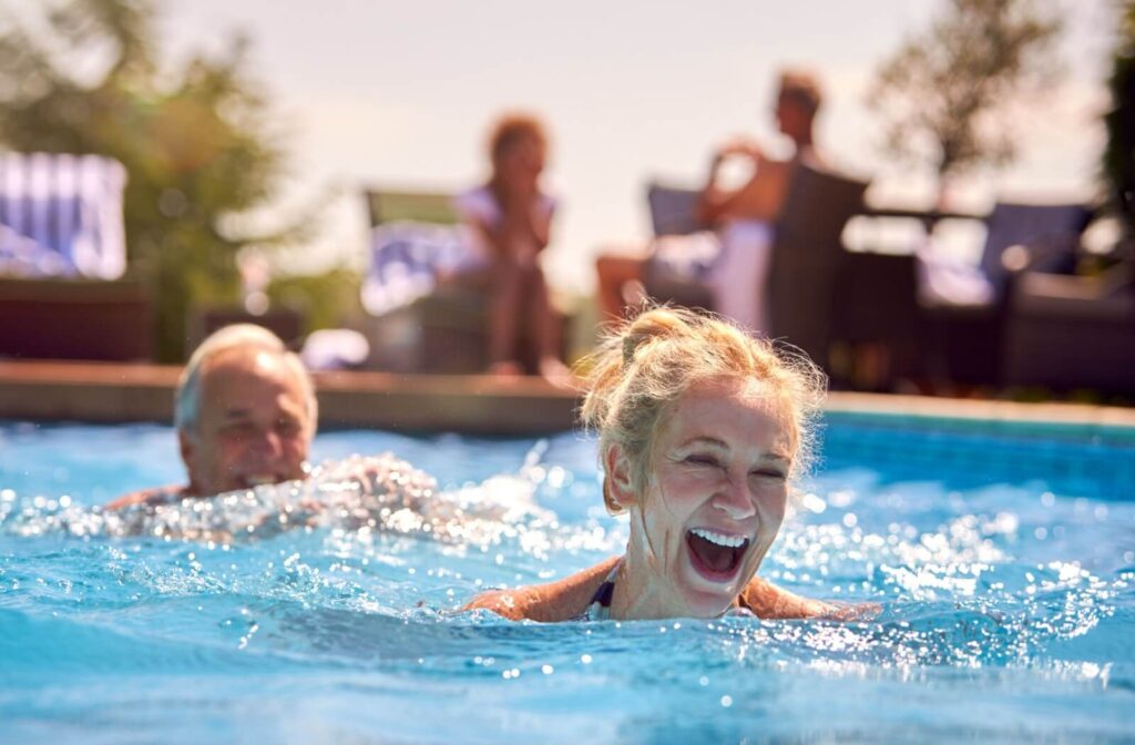 Smiling older adult swimming in outdoor pool with others relaxing nearby on a sunny summer day