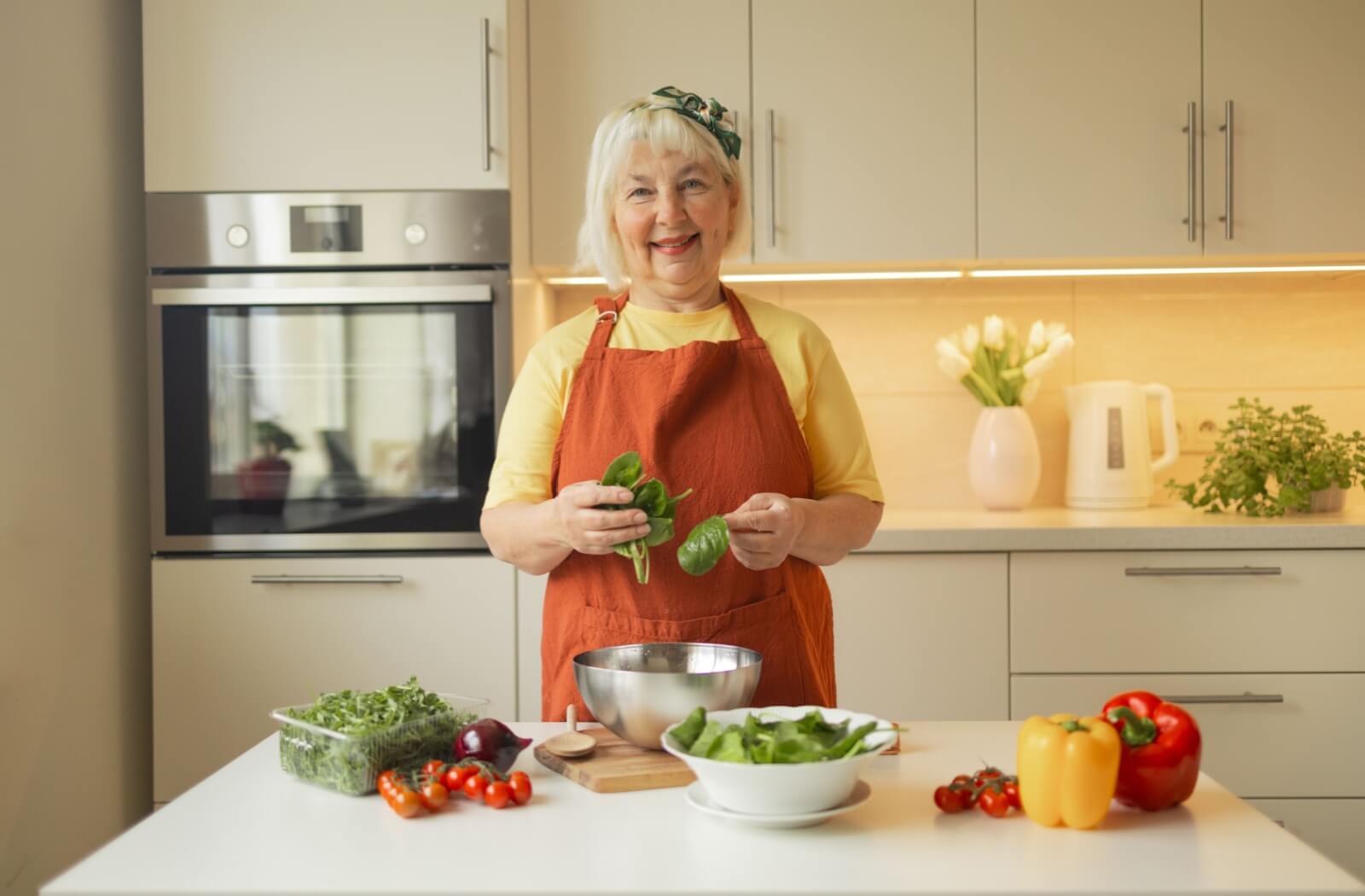 Smiling senior in orange apron holding spinach in bright kitchen with fresh vegetables