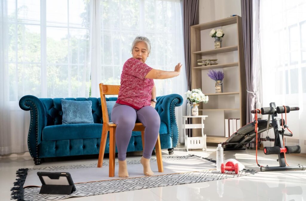 A senior sitting on a chair in the middle of their living room follows along to video instructions for chair yoga positions