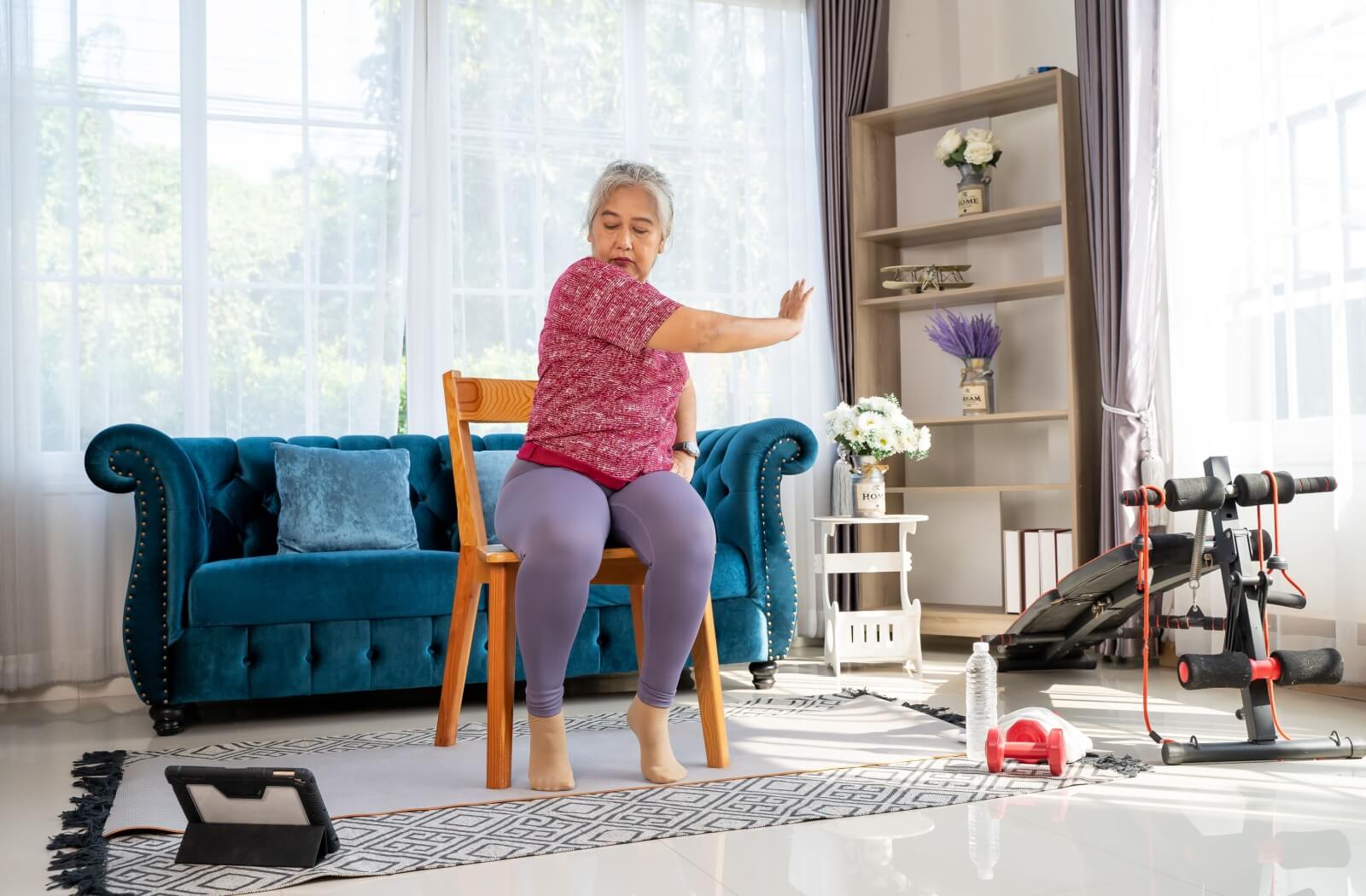 A senior sitting on a chair in the middle of their living room follows along to video instructions for chair yoga positions