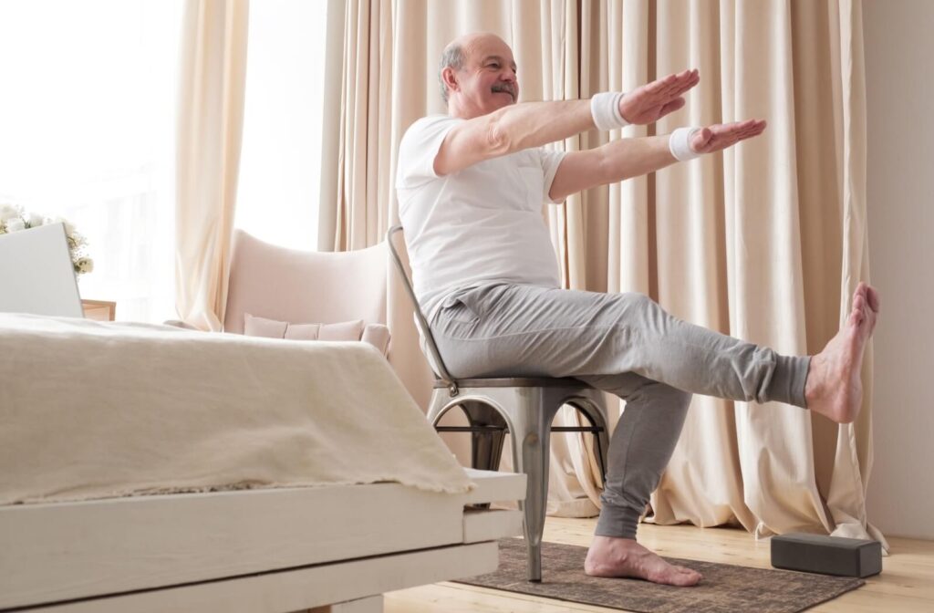 A senior sitting on a chair in their bedroom performs a leg and arm raise yoga position to improve balance