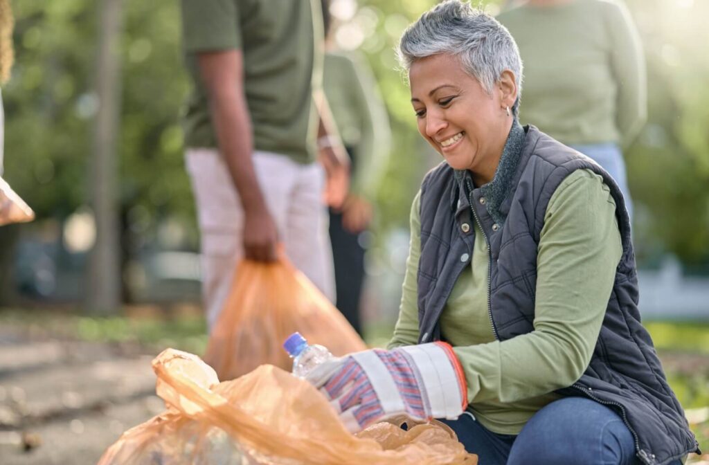 A senior wearing gloves bends down to pick up trash from a park as part of a volunteer community clean-up effort